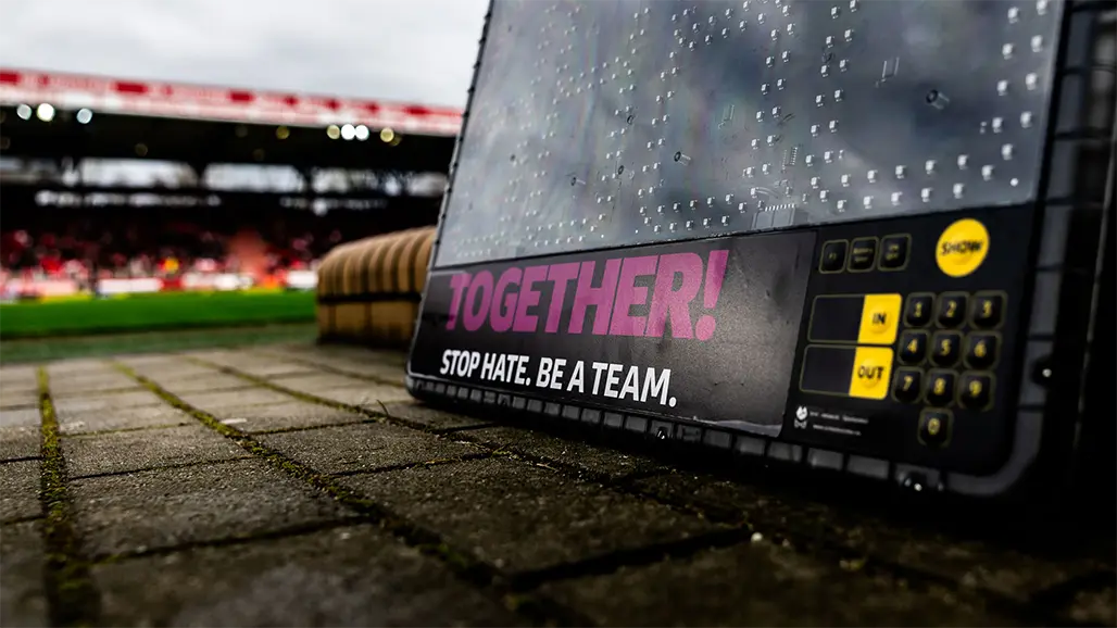 The image shows a product display with a promotional banner or tarpaulin over the background. It appears to be taken on an outdoor sports court in what seems to be wet or damp weather conditions, as suggested by the reflections on the tarp and puddles on the ground. The sign on top has white text that might be associated with an event or team name. In the foreground, there's a piece of equipment which looks like a portable electronic scoreboard or a display unit for viewing scores or other game-related information, given the presence of various control buttons. There is also a logo and tagline on the device, although the text resolution is low, making it difficult to read clearly. The sports court has a surface that could be used for activities like football or rugby, and there's some signage in the distant background with what looks like more writing that might provide further context about the location or event.