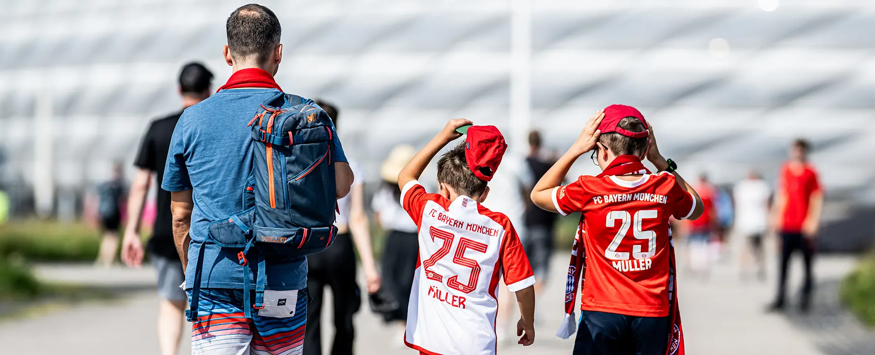 This is an image of a group of people, possibly a family or team members walking together outdoors. They appear to be at an event, indicated by the banner in the background which might refer to a sports or recreational activity. The central figure is holding up his hand for a salute or wave, while others are looking ahead. Everyone seems dressed appropriately for the weather or the occasion, and one individual has a backpack slung over their shoulder. The setting looks like an open-air area with paved paths, suggesting it could be within a park, sports complex, or another public space.