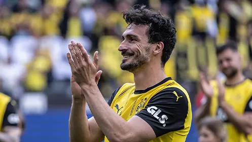 You've shared an image of a man, likely a soccer player, who appears to be standing on a field or a stage during an event. He is looking towards the camera and has a proud or content expression on his face. The background seems to be filled with spectators, indicating that this might be a professional sporting event. The man is clapping, suggesting that he might have scored a goal or won a competition for his team. His body language indicates celebratory joy and perhaps exhaustion from the exertion of playing in matches like the World Cup of Soccer.