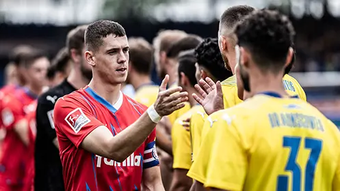 This is an image of a person who appears to be a soccer player. The individual is standing in what looks like a locker room or the sidelines, wearing a red and white striped top, which is often the color scheme for a national team or an international club, suggesting they may be at a professional level. There are hands on shoulders and head gestures with people in close proximity, indicating some form of interaction or communication between them. The background shows another player who seems to be looking towards the standing individual.