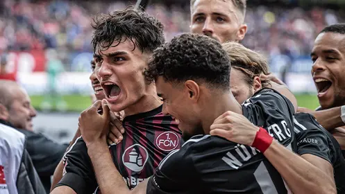 The image features a group of soccer players in celebration following the conclusion of a game. One player is holding up something, possibly a trophy or a cup, and appears to be cheering with his teammates around him. The setting suggests that this could be a post-match scene from a soccer stadium as indicated by the seating structure visible in the background behind them.