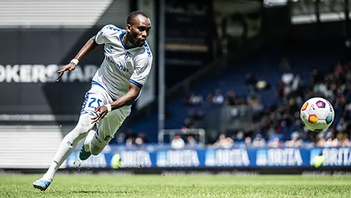 In the image, there is a soccer player in the act of kicking the ball. He appears to be in the midst of a run and has just made contact with the ball using his laces on his left foot. The player is wearing a white kit with blue accents, which is not unusual for away teams due to sponsorship, as their colors may differ from those of their home team. The stadium background suggests this is a professional soccer match due to the lighting, stand design, and the image captures a pivotal moment that is likely integral to the game's outcome. The focus on the player during this action shot highlights the dynamic nature of sports like soccer.