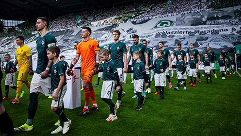 The photo depicts a group of soccer players walking together across what appears to be a grassy soccer field. They are dressed in matching kits, with the main color being green and some white elements, which might suggest they belong to a single team. In the background, there is a large flag bearing the insignia of what looks like a sports club, indicating that this could be at the venue of a professional soccer match. The environment includes stadium seating on either side of what seems to be the field, and the lighting conditions suggest it might be evening or that the venue is indoors with artificial light.