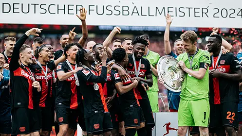 The image shows a group of soccer players celebrating. They appear to be holding a trophy, which suggests that they may have won or achieved something significant during their sporting endeavors. The setting seems to be outdoors with flags and banners indicating a public event or location, likely related to the sport celebrated by the players. There's also an accompanying watermark in German that likely refers to the brand sponsoring the event.