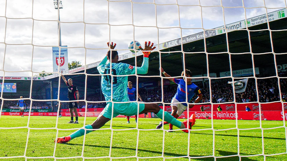 Torwart Tyler Doğan streckt sich im türkisfarbenen Holstein Kiel Trikot nach einem Schuss, während der Ball direkt vor seinen Händen auftaucht und Spieler im Hintergrund reagieren. (Foto)