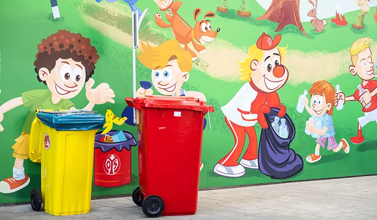 At the FSV Mainz 05 stadium, a yellow and a red rubbish bin stand in front of a painted wall depicting a clean-up operation in the forest. (Photo)