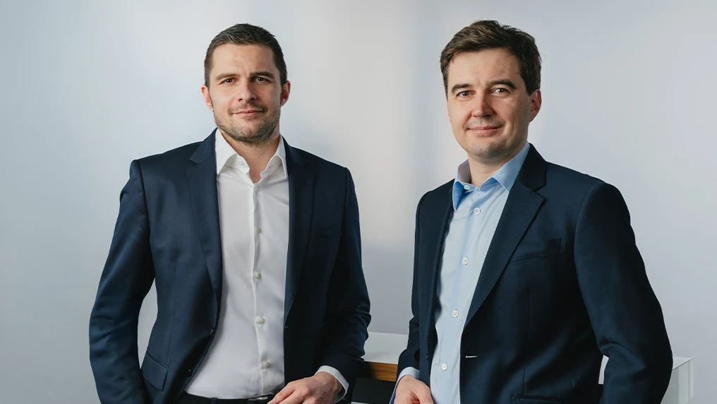 Two brunette men wearing dark blue suits, smiling into the camera. (Photo)