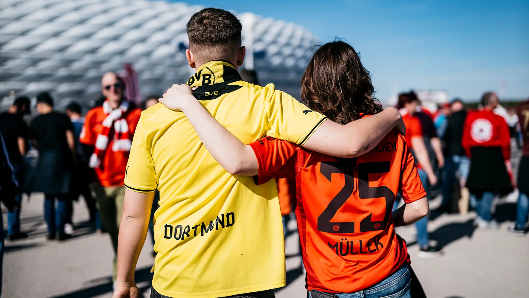A man in a BVB jersey and a woman in an FC Bayern jersey are standing in front of a stadium. They put their arms around each other's shoulders. (Photo)